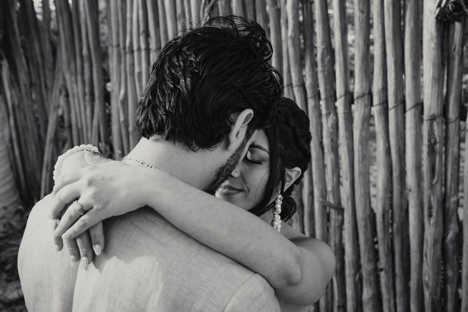 Bride and groom share emotional embrace near rustic bamboo fence in black and white.