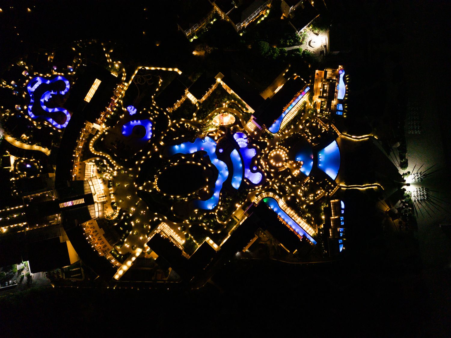 Aerial night view of a resort complex with golden lights and vivid blue pools forming a symmetrical pattern.