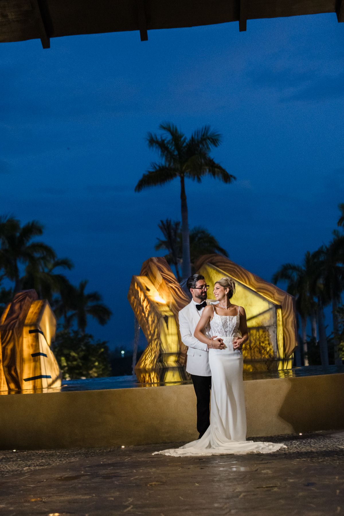Bride and groom pose elegantly at twilight near illuminated resort with palm trees in tropical wedding setting.