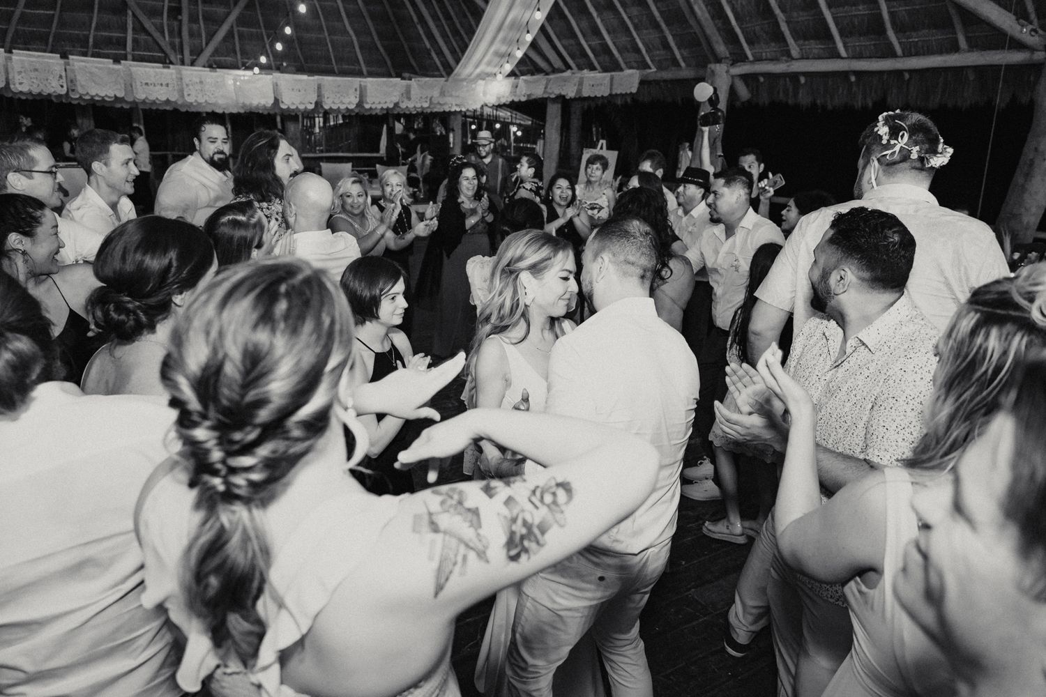 Bride and groom surrounded by cheering guests on packed wedding reception dance floor in black and white.