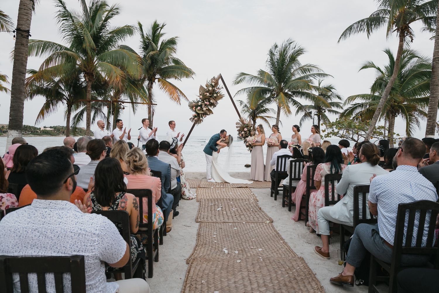 Guests seated at elegant outdoor beach wedding ceremony with palm trees and floral arch altar.