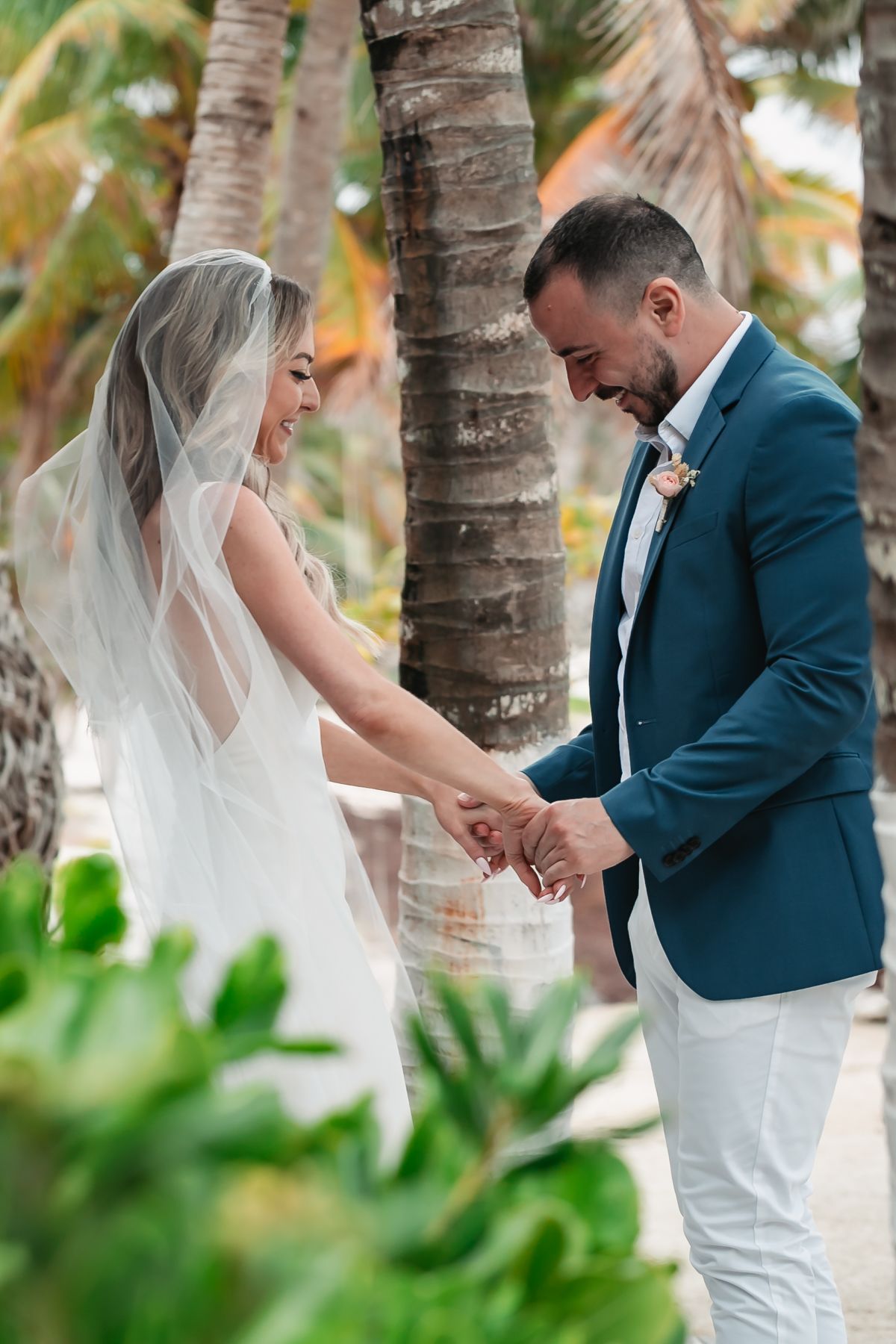 Bride and groom holding hands and smiling among lush palm trees at a tropical beach wedding.