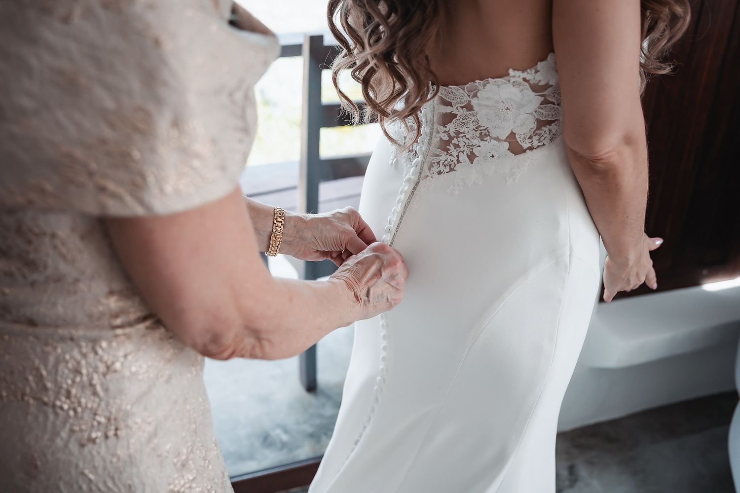 A woman helps a bride zip up her elegant white lace wedding dress from behind near a window.