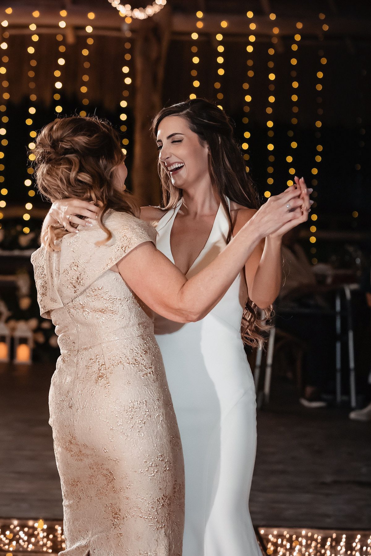 Bride in white gown dancing with guest in lace dress at elegant wedding reception with warm bokeh lights.