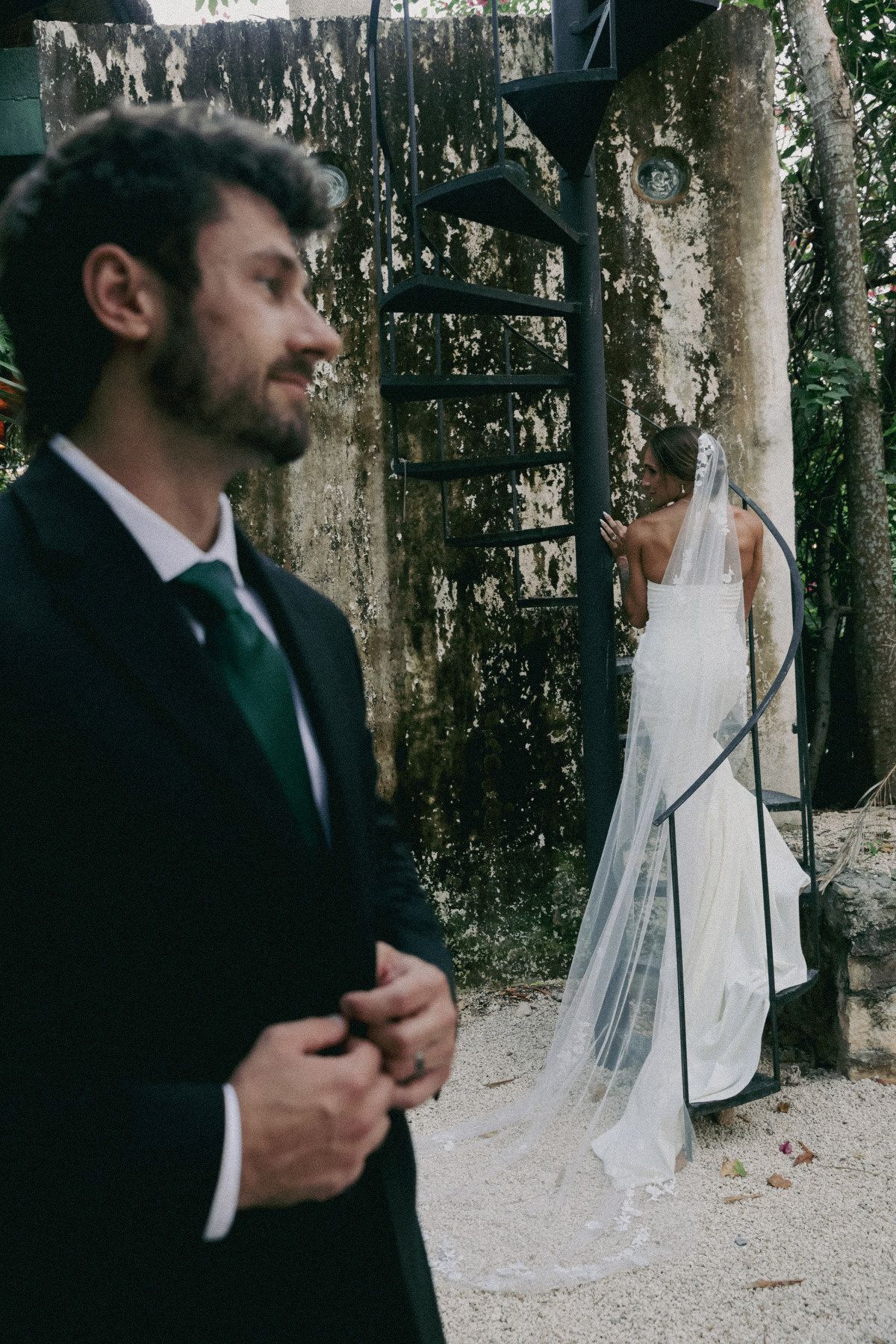 Groom in black tuxedo smiles as bride in white gown and veil stands by spiral staircase outdoors.