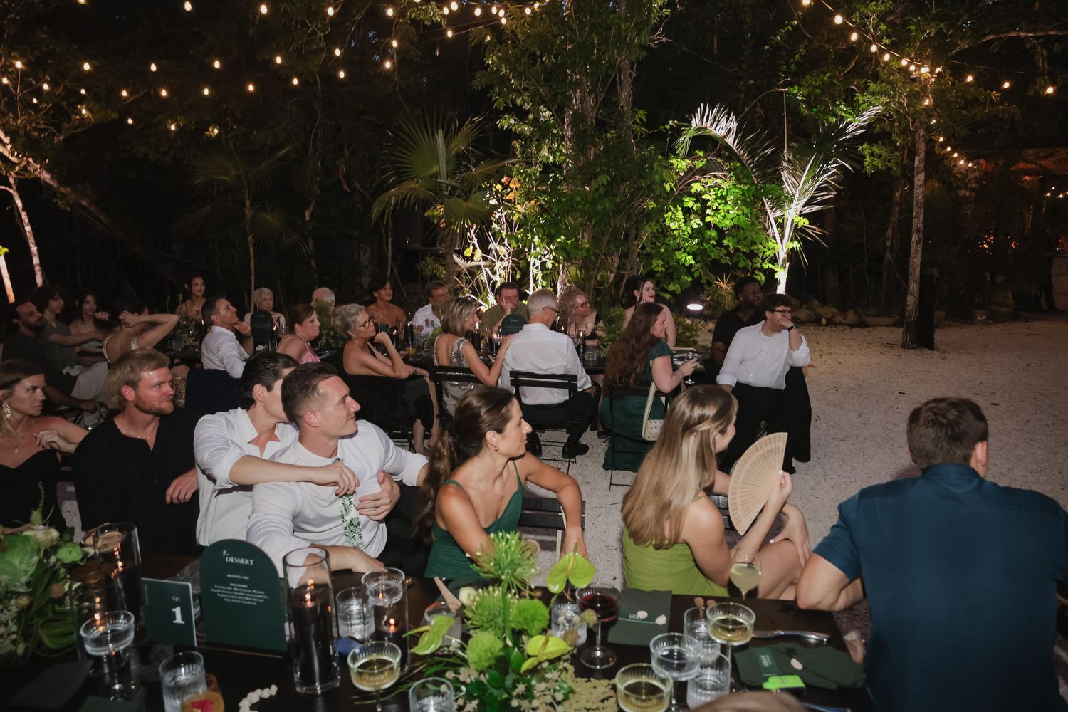 Guests gathered at an outdoor wedding reception under string lights surrounded by tropical trees at night.