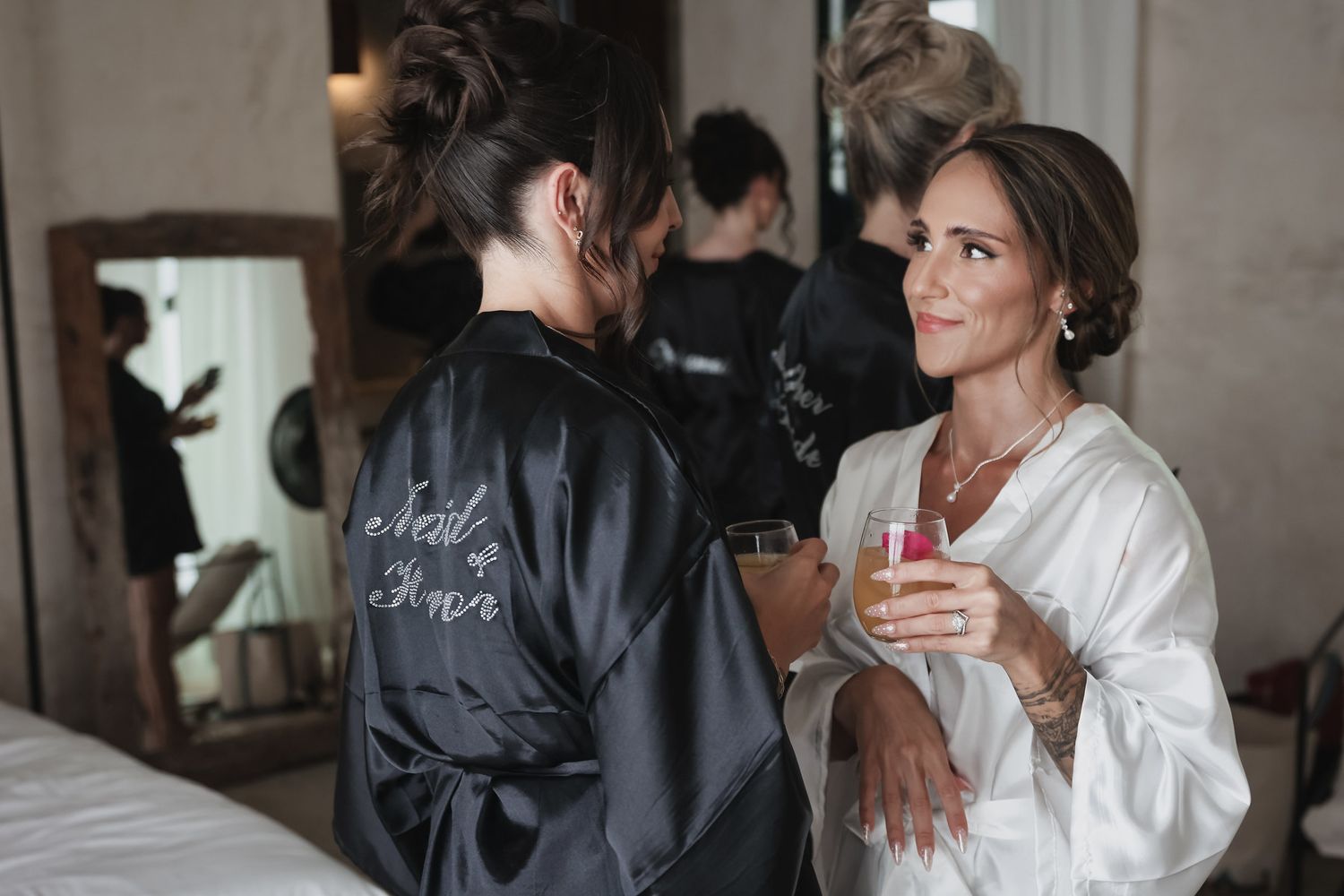 Two women in satin robes, one labeled bridesmaid, share a toast with champagne during bridal preparations.
