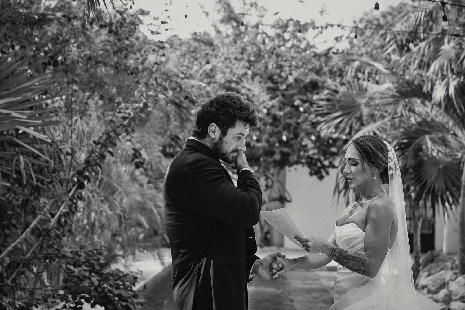 Black and white photo of emotional groom wiping tears as bride reads wedding vows outdoors.