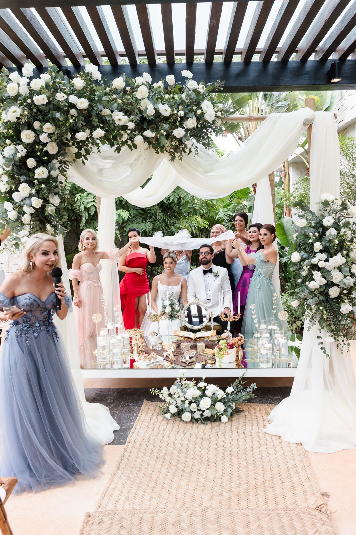 Wedding ceremony with bride and groom under floral arch, surrounded by bridesmaids in colorful gowns outdoors.