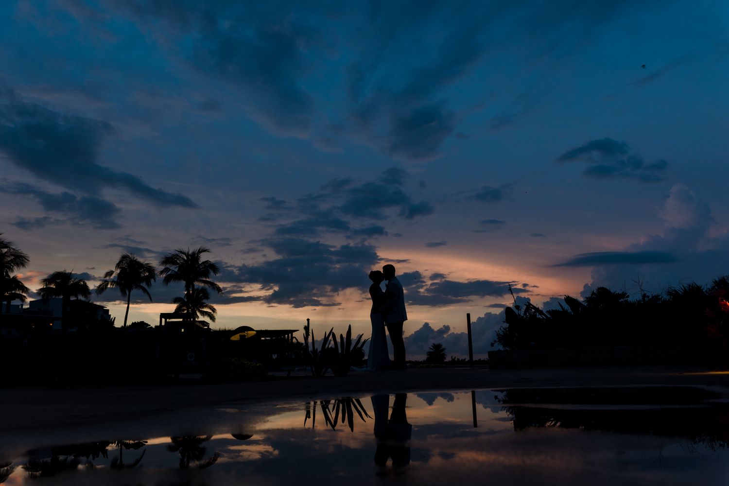 Silhouette of a couple embracing at sunset reflected in water with palm trees and dramatic blue sky.