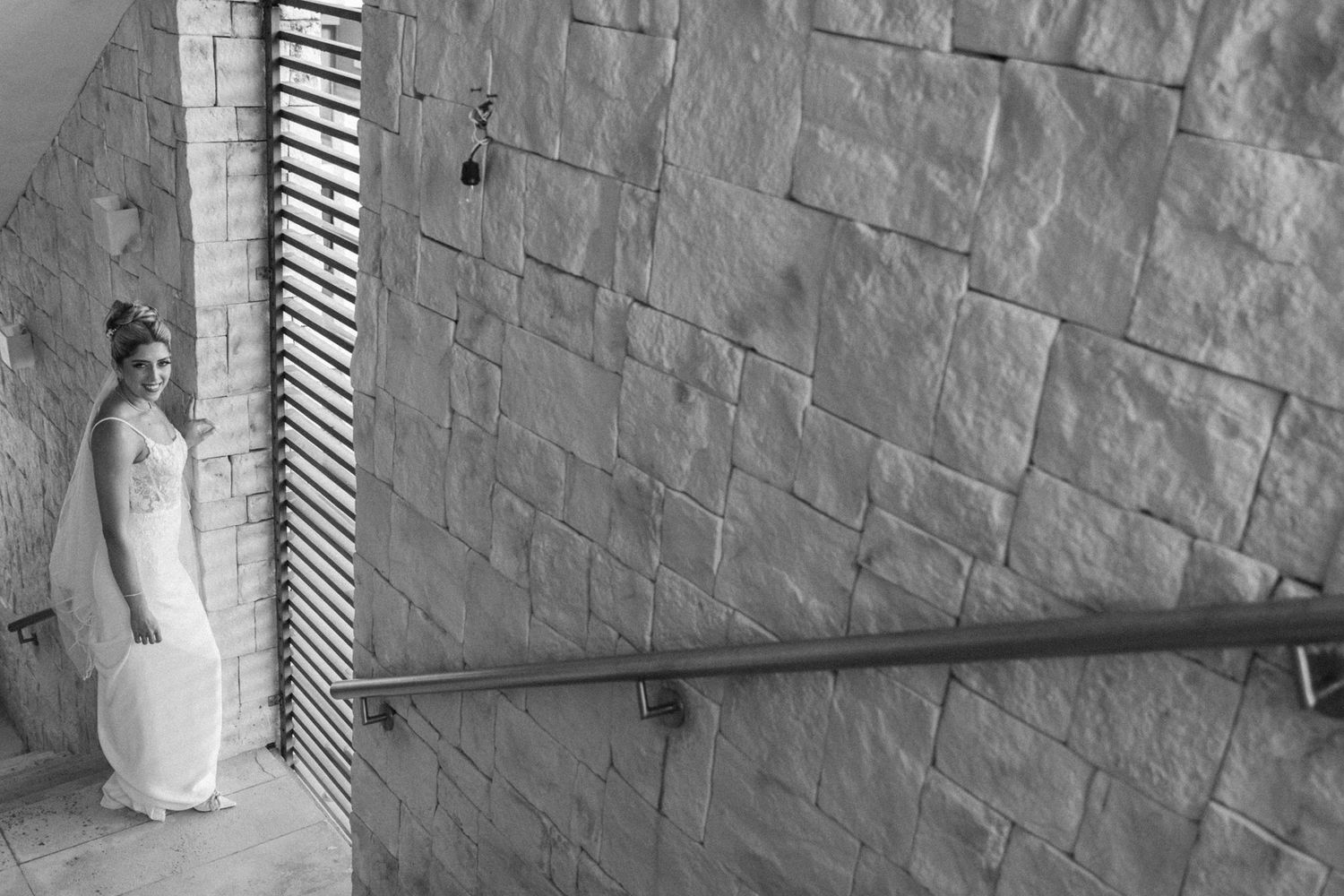 Bride in white gown standing by stone wall with handrail, black and white wedding portrait photography.