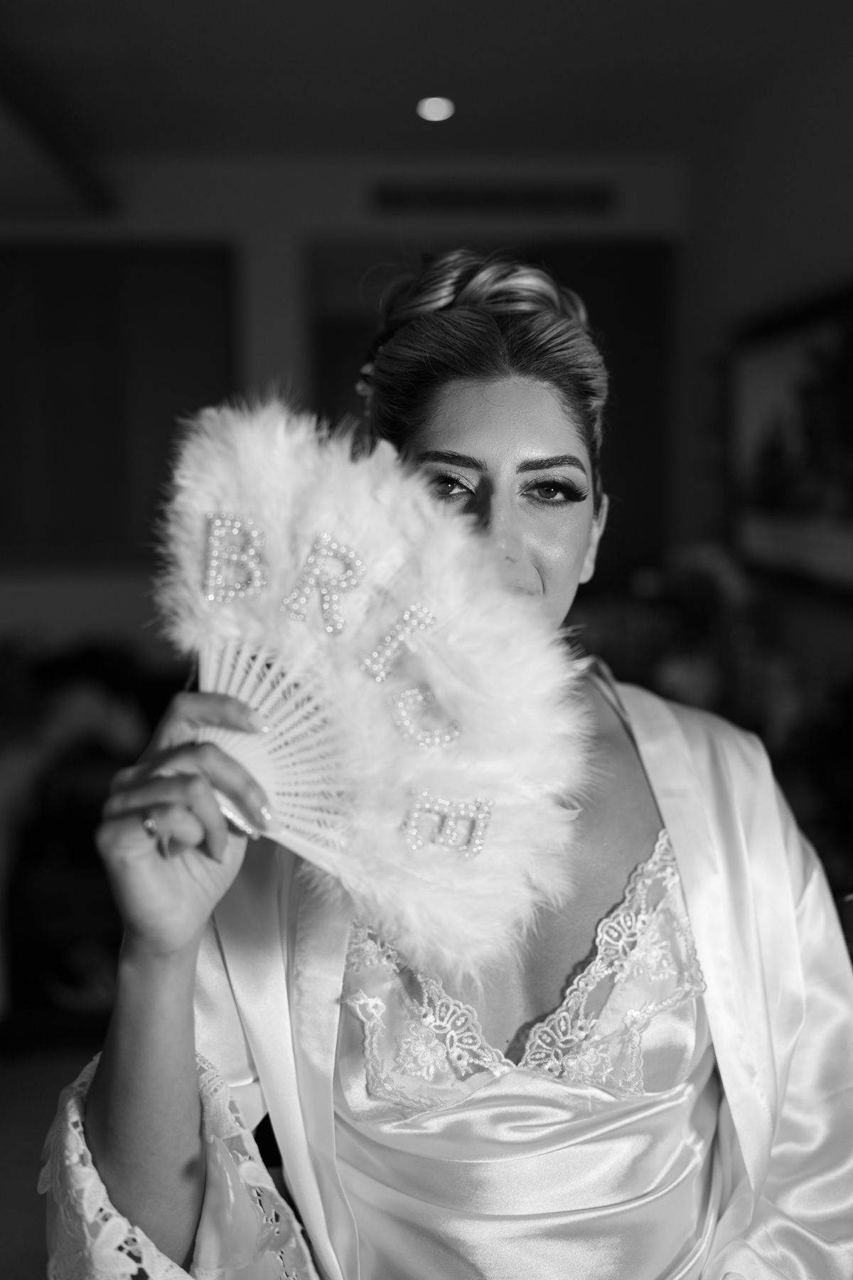 Black and white photo of a bride holding a feathered fan with BRIDE written on it, wearing a satin robe.