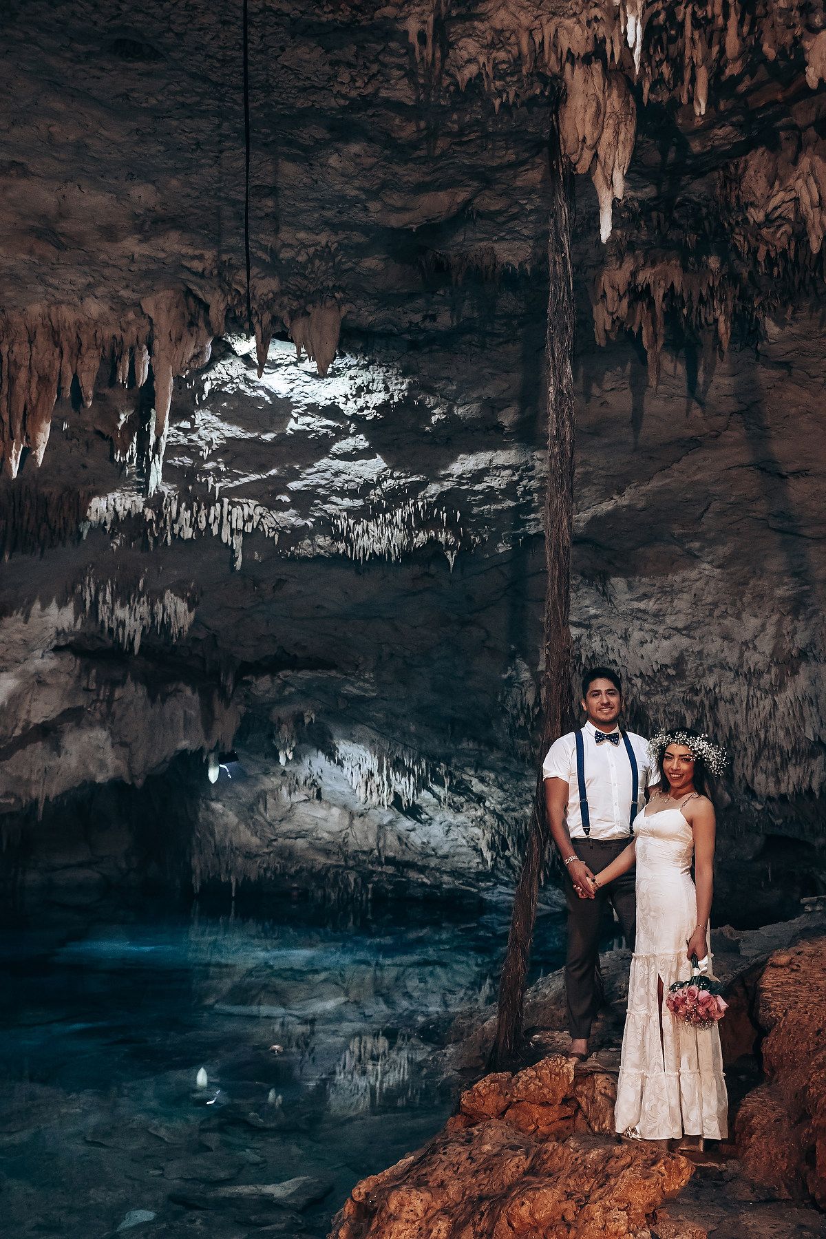 A romantic photoshoot in a dimly lit cave with dramatic stalactites and turquoise water in the background.