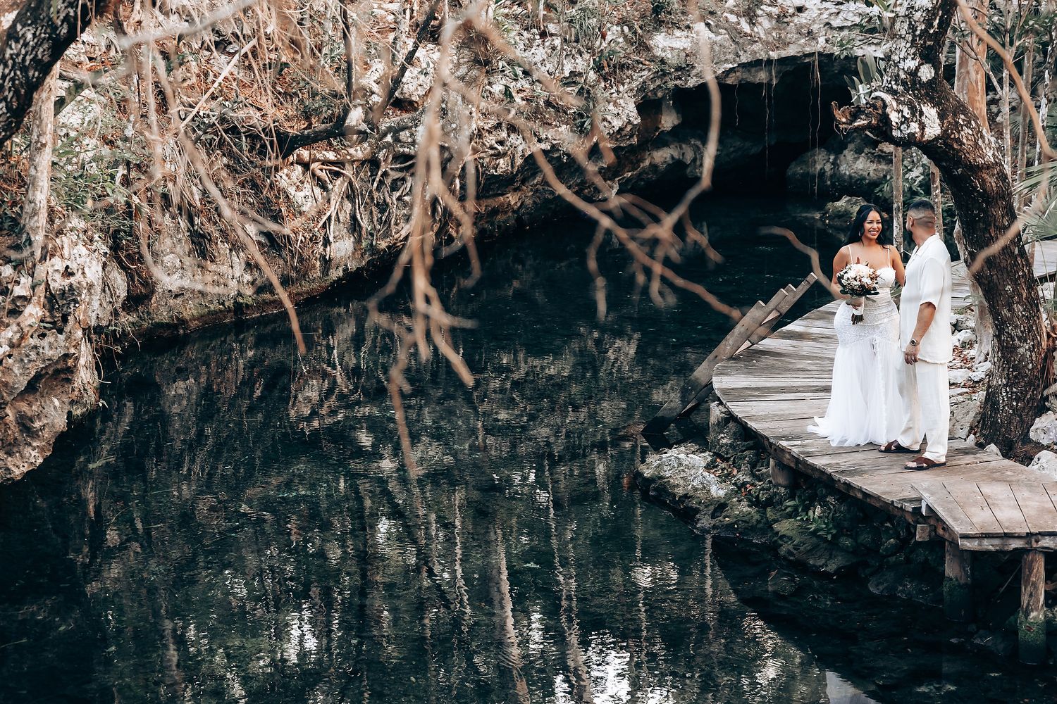A couple stands on a wooden dock over a serene cenote surrounded by forest.