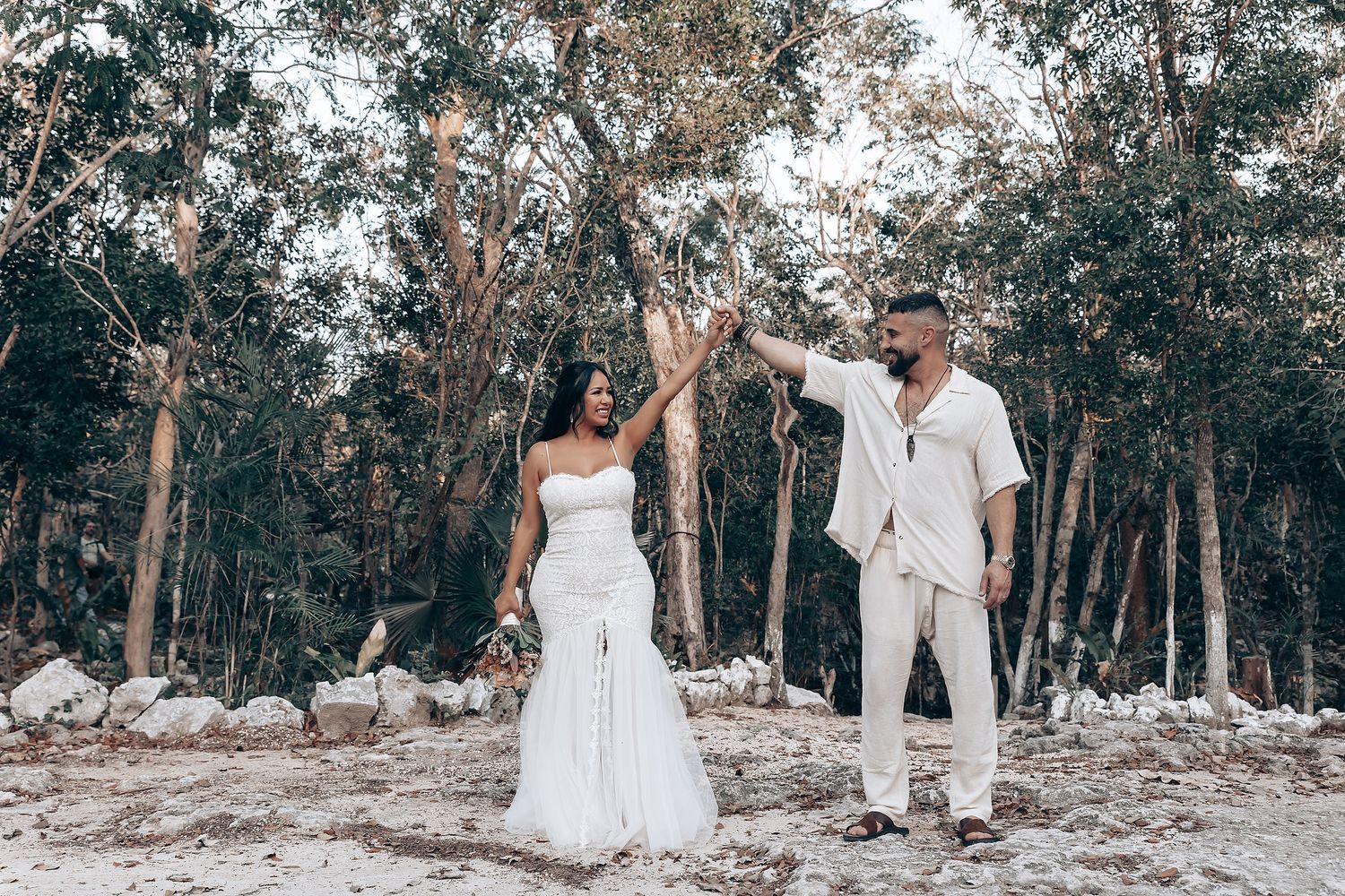 A couple celebrates their wedding day in an outdoor forest setting wearing matching white outfits.