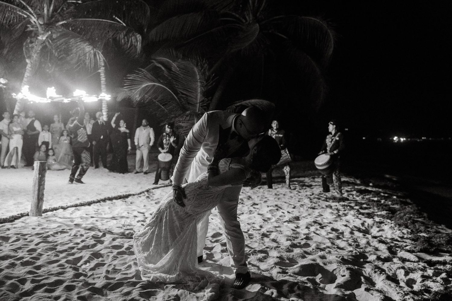 Couple shares romantic dip kiss on sandy beach surrounded by wedding guests at night celebration.