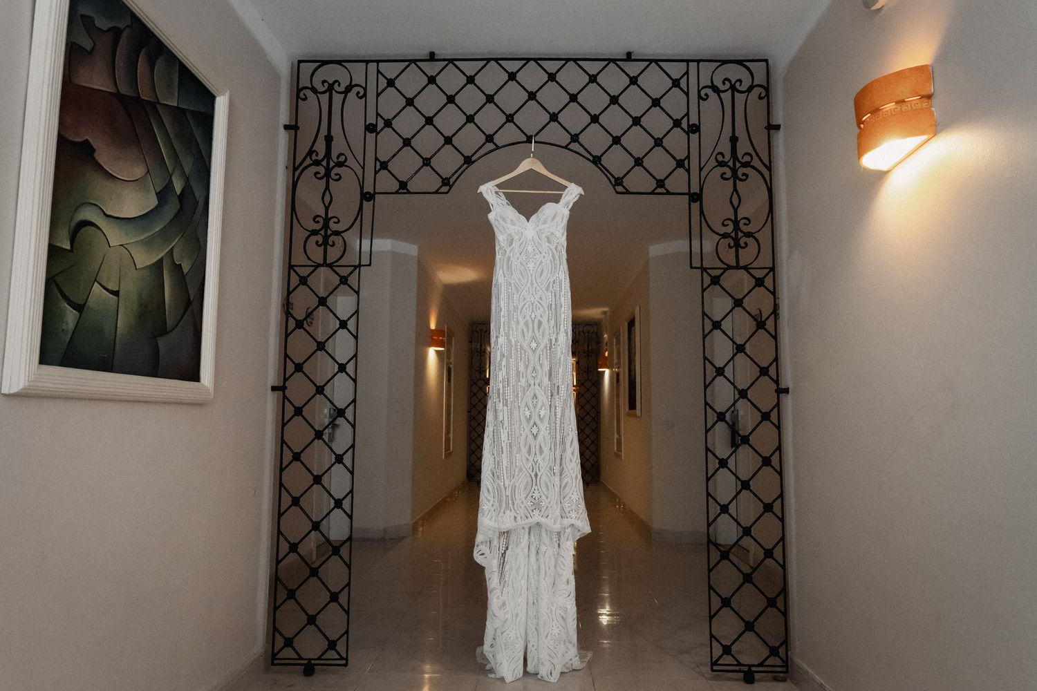 A white lace wedding dress hanging on an ornate wrought iron gate in an elegant hallway with warm lighting.