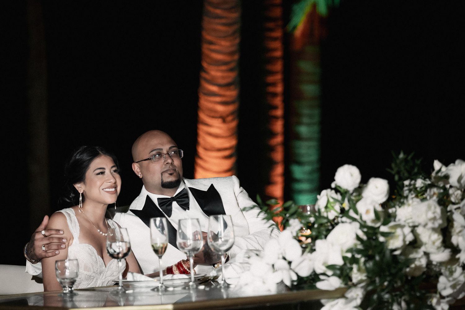 Bride and groom smiling at sweetheart table with white floral arrangements at reception.