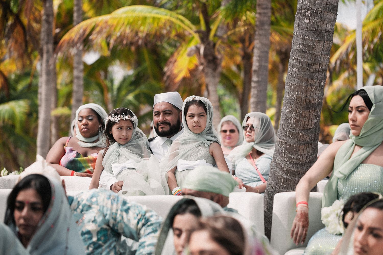 Wedding guests in white and green head coverings seated outdoors under palm trees at Sikh ceremony.