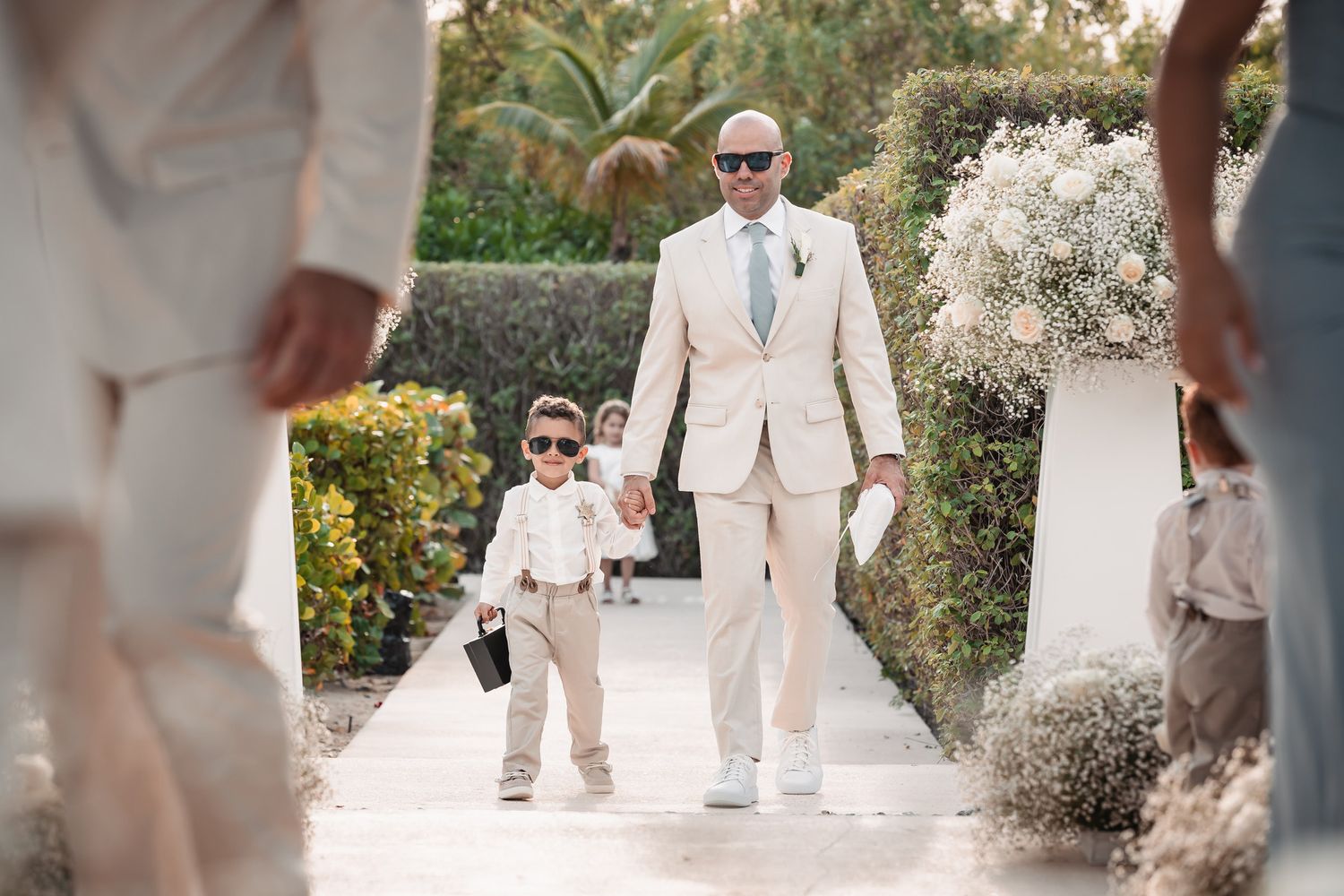 Dapper man in beige suit walking with a mini ring bearer in matching sunglasses down the aisle.