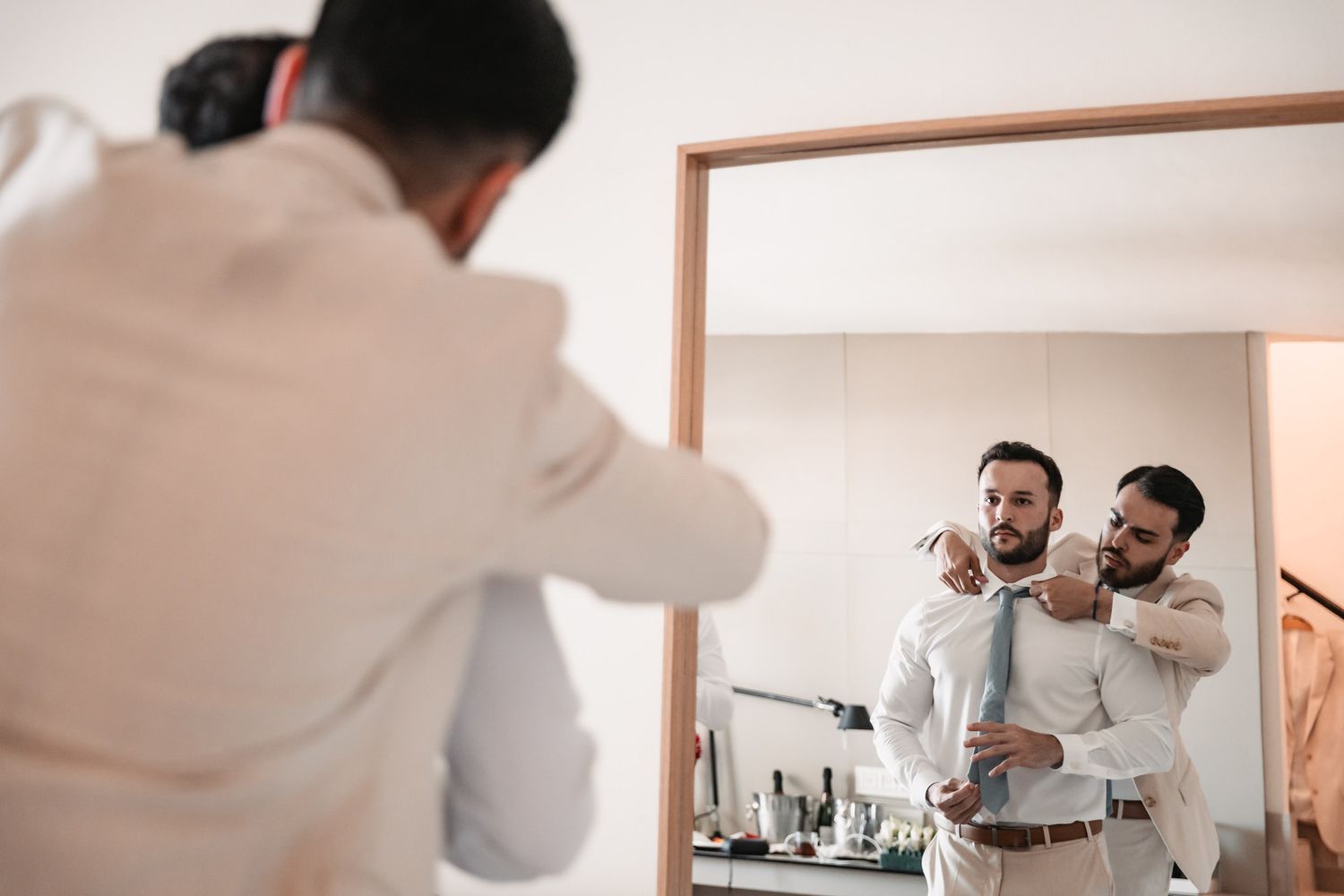 Two men in formal attire help each other with ties while preparing for a wedding, reflected in a large mirror.