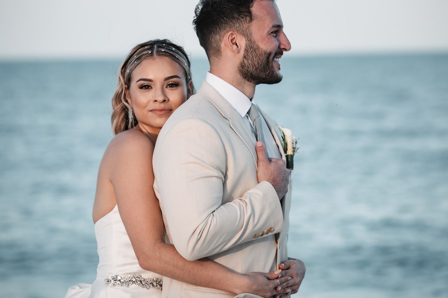 Bride and groom embracing back-to-back on beach in wedding attire at sunset.
