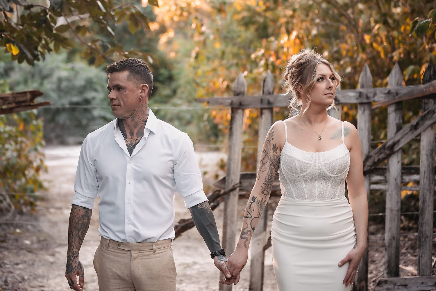 Tattooed couple in wedding attire holding hands on a rustic dirt path surrounded by autumn trees.