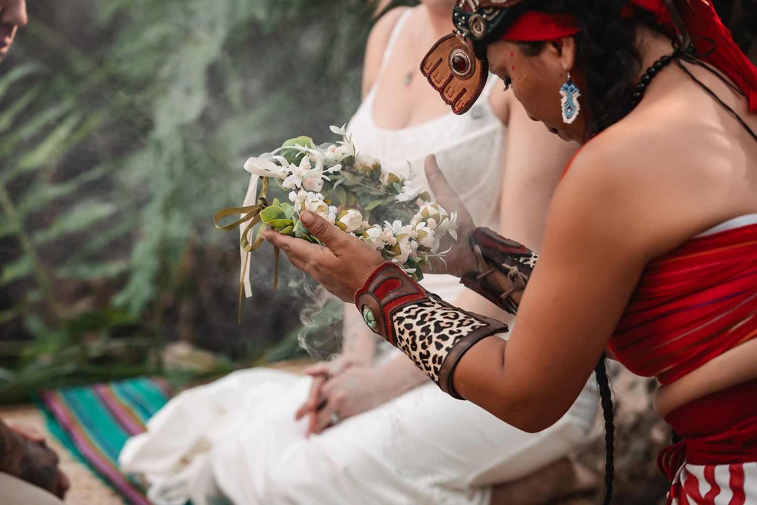 Indigenous ceremonial ritual with woman in red holding white flower wreath, wearing leopard print bracelet outdoors.