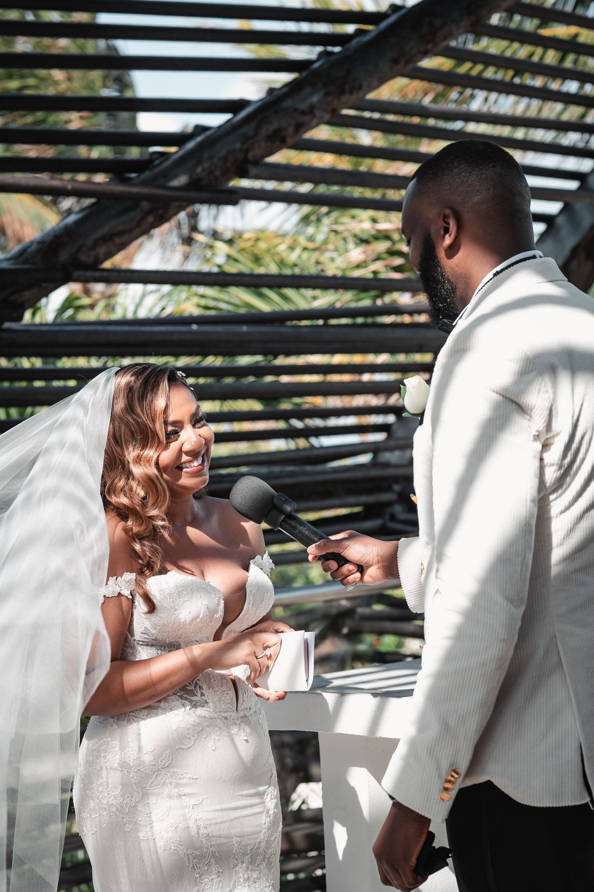 Radiant bride reading vows to groom in white suit under outdoor wooden pergola.