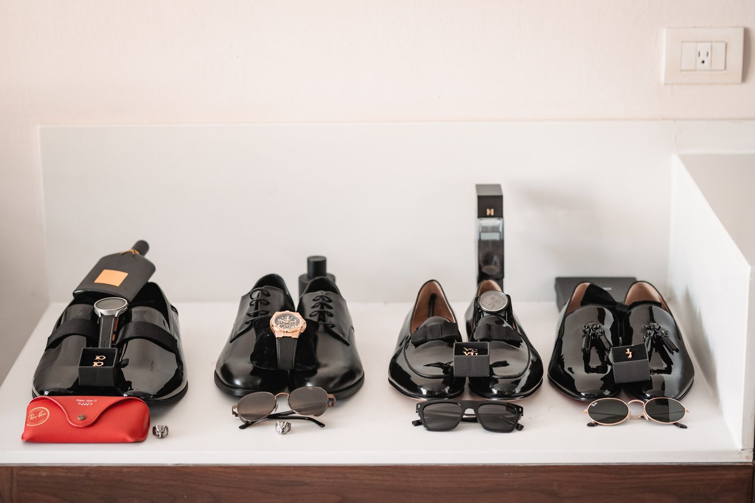 Four pairs of black dress shoes displayed on a shelf with watches, sunglasses, and accessories for a formal occasion.