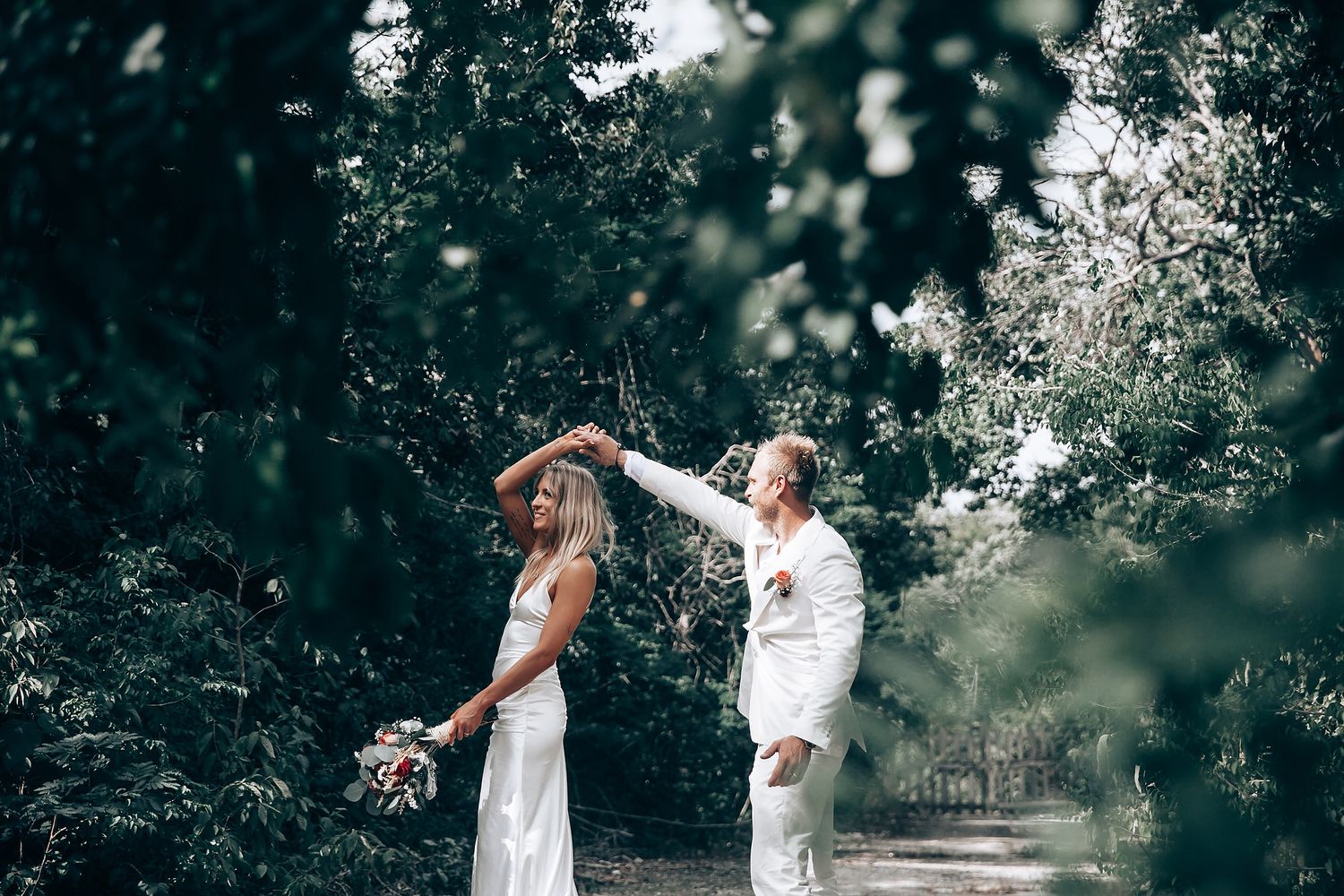 A joyful couple in white clothing dance together in a sunlit forest setting.