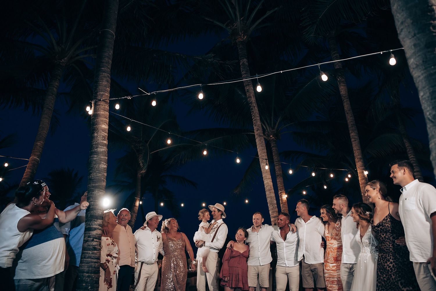 Evening wedding reception with string lights illuminating palm trees and dancing guests.