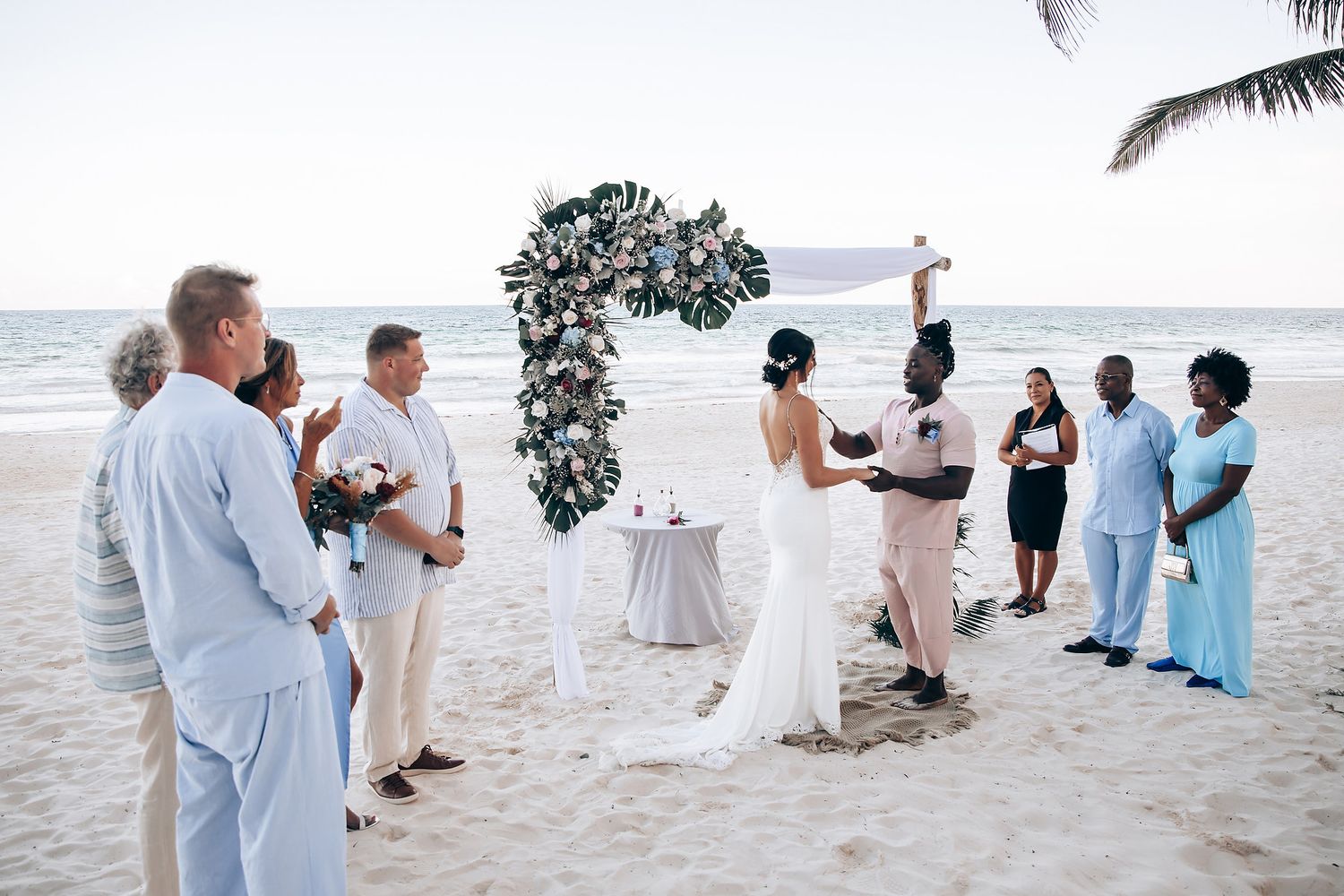 The image shows a bride and groom standing together on a beach, surrounded by their wedding party.