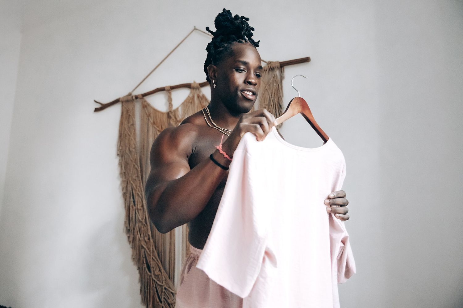 A shirtless man with dreadlocks is holding a white towel in front of a white wall.