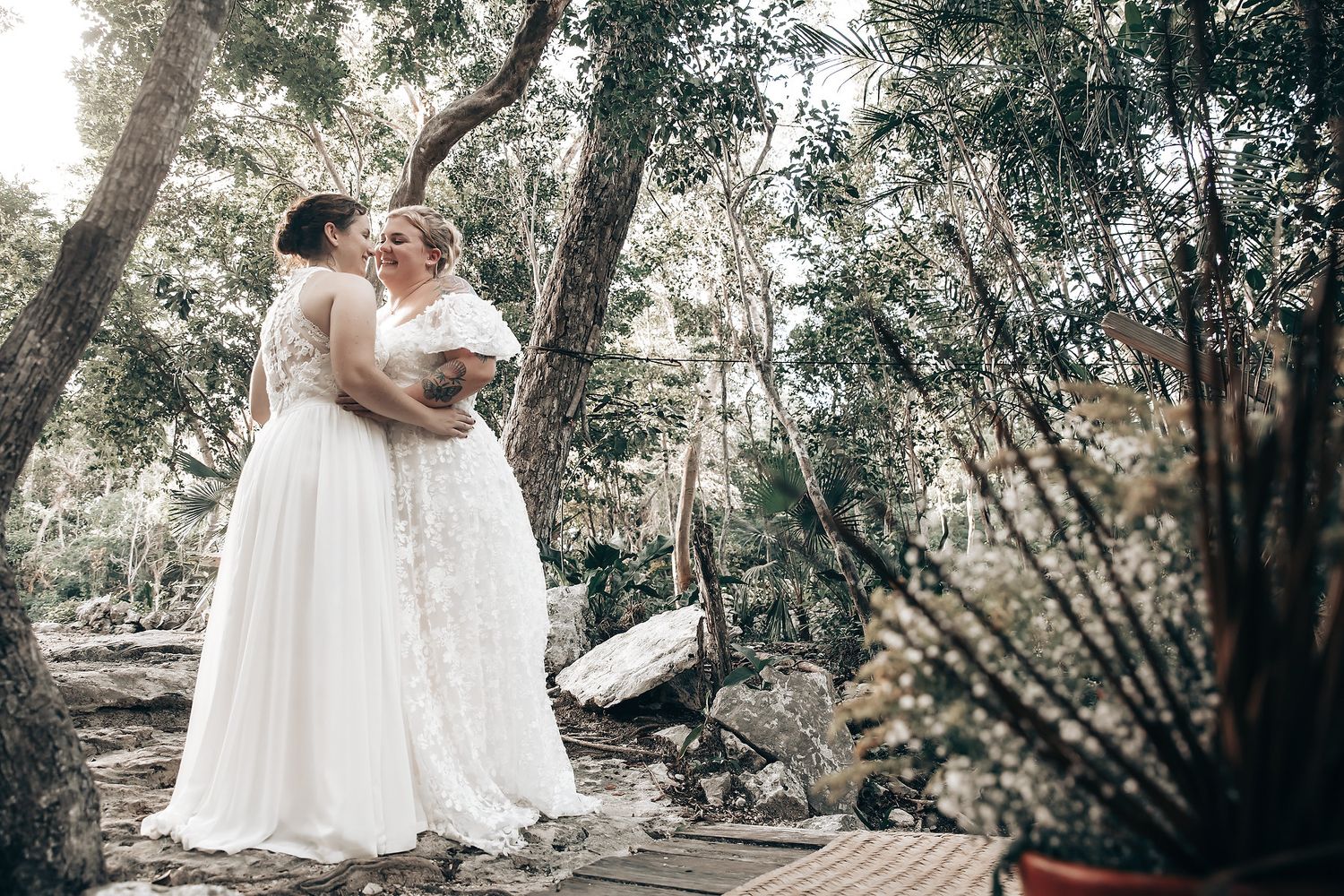 Two women in white wedding dresses standing on a wooden platform in a forest with trees and foliage in the background.