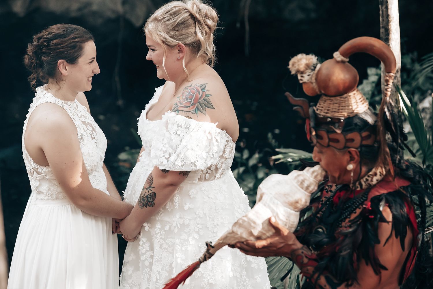 Two brides in white wedding dresses standing together with a person in traditional indigenous attire.