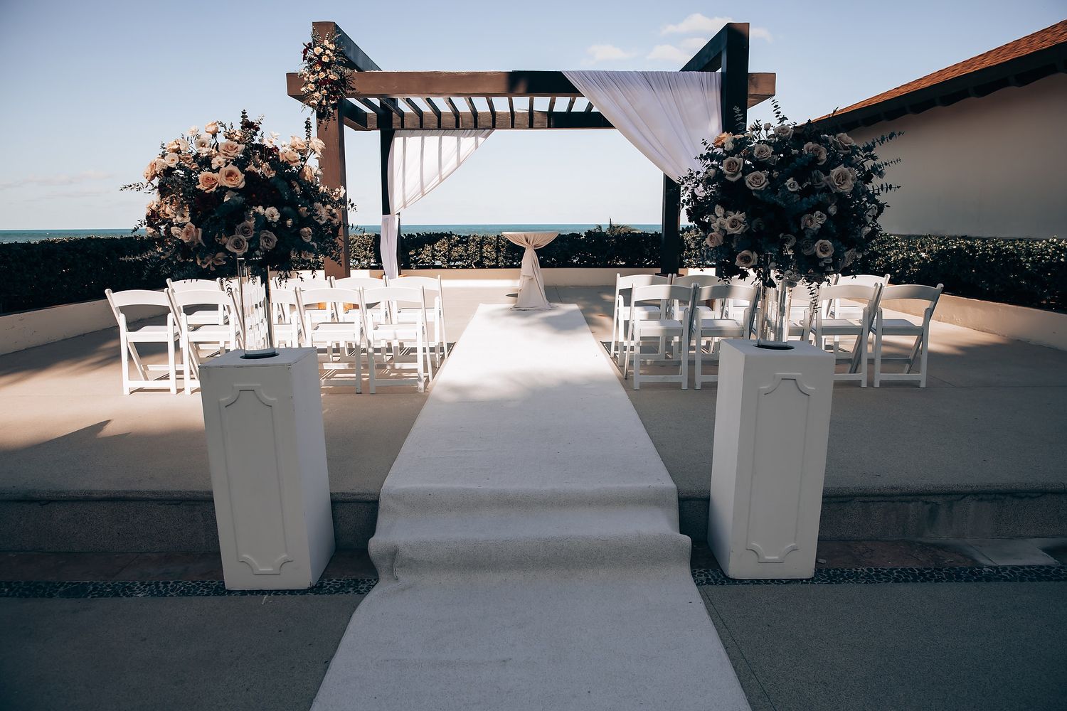 A wedding ceremony setup with an arch, white chairs, and a white aisle leading to the ocean.
