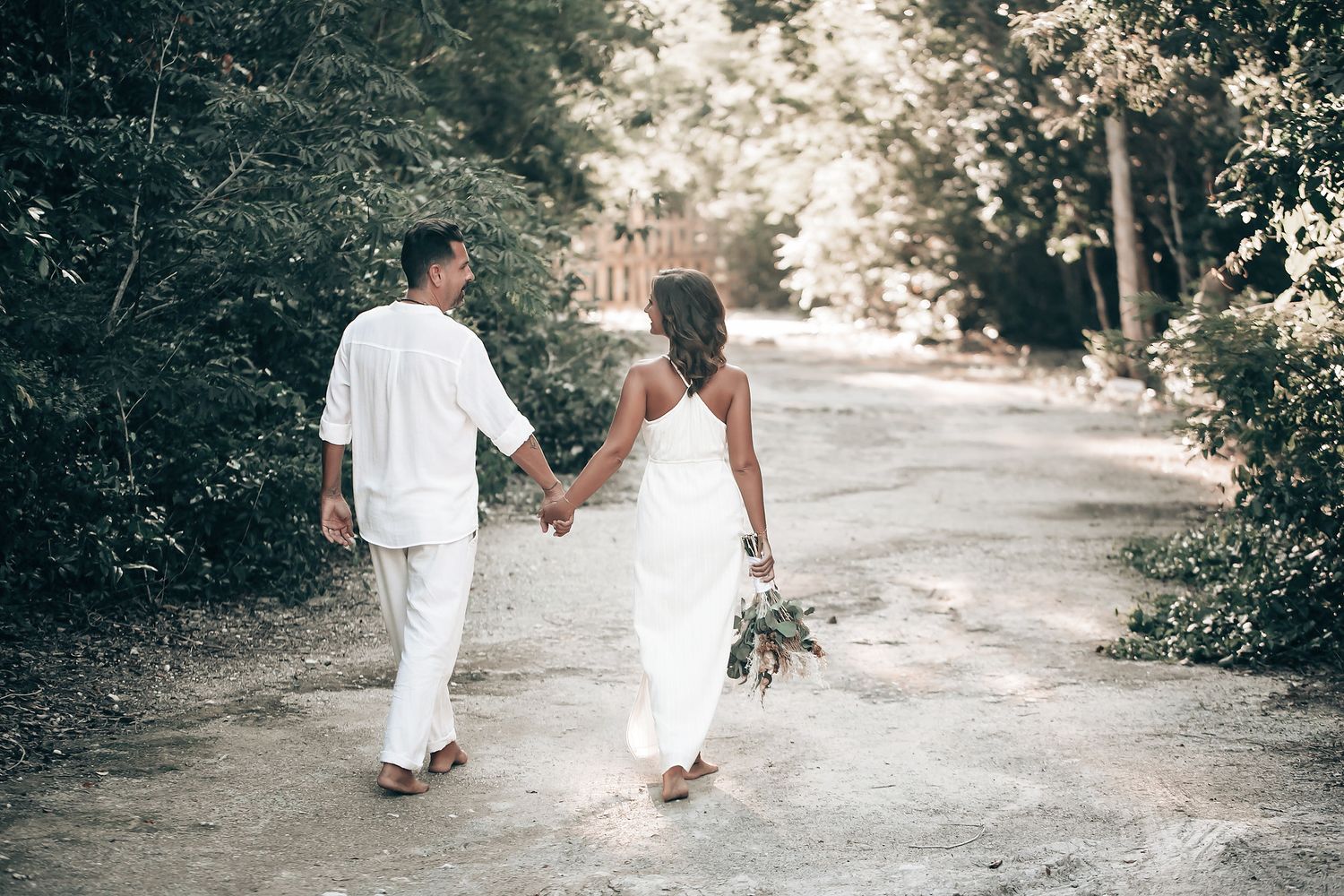 A couple dressed in white clothing walk hand-in-hand down a path in a wooded area.