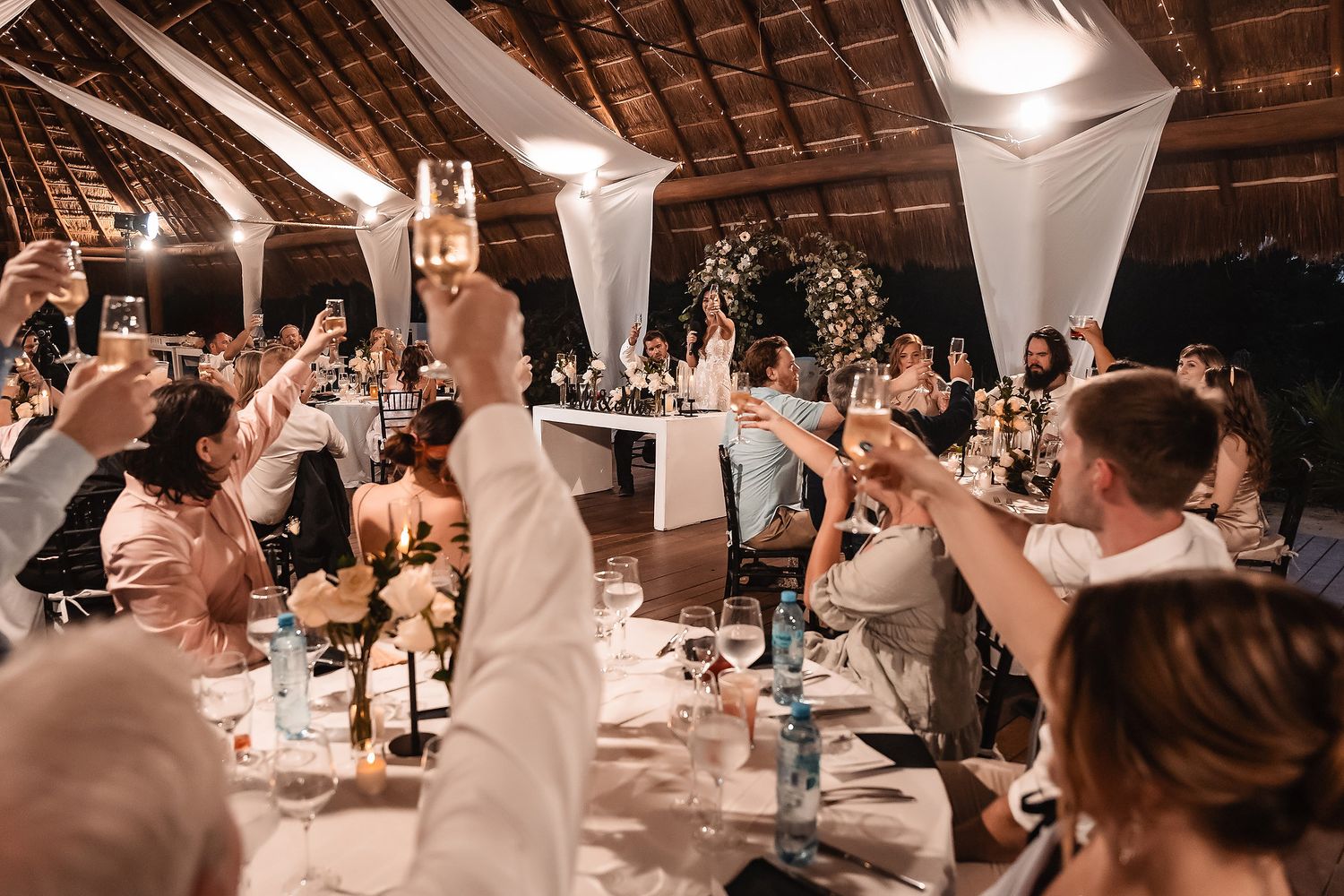 Guests raising champagne glasses in a toast at an elegant outdoor wedding reception under a thatched roof.