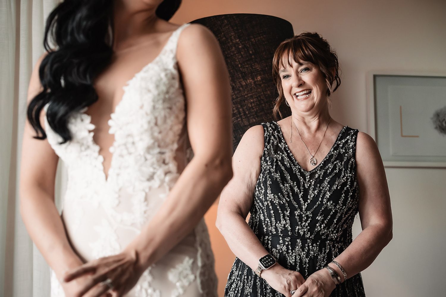 Emotional mother smiling proudly as bride stands before her in a lace wedding gown.