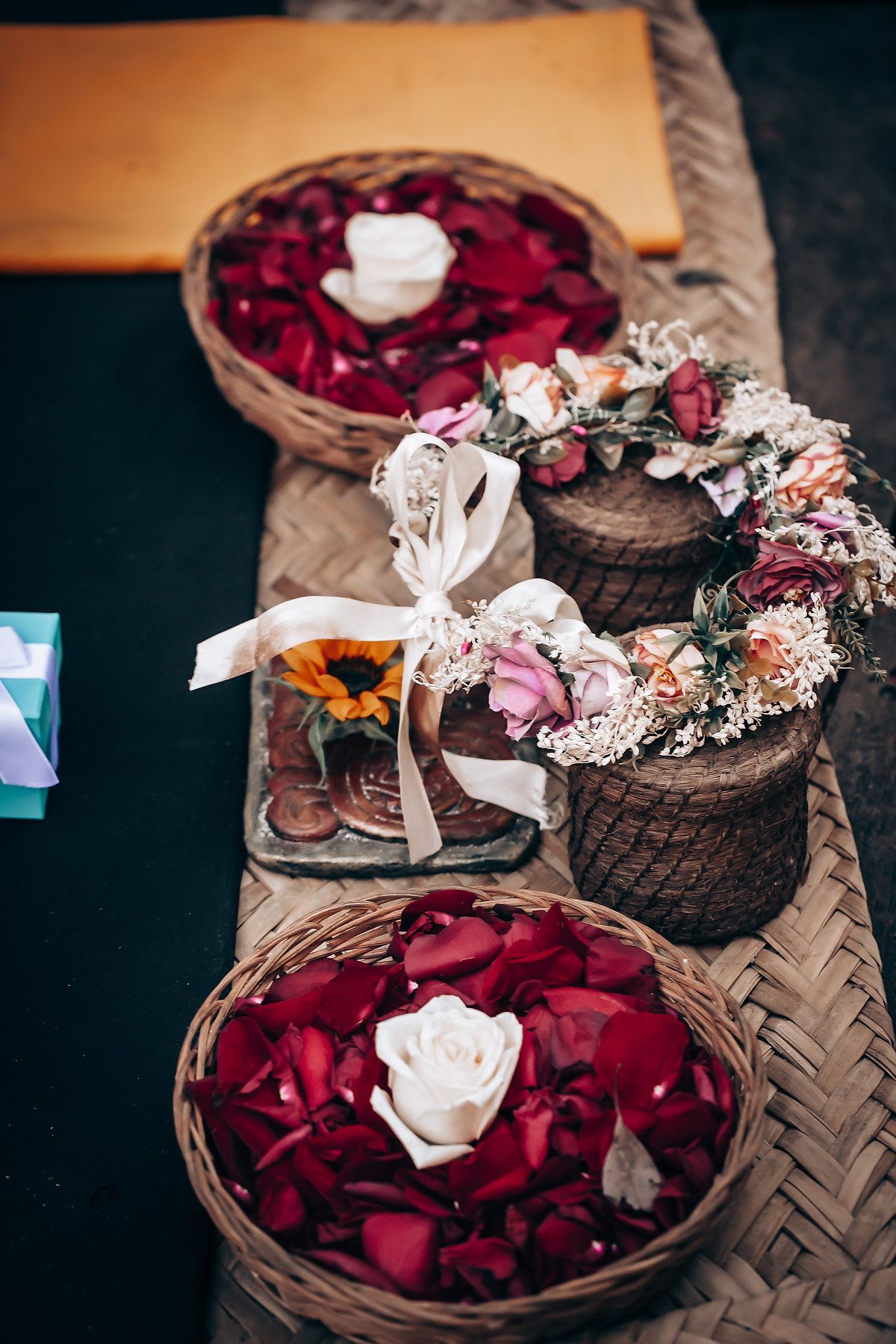 Romantic wedding decor with red rose petals in woven baskets, floral wreath, and decorated glass jar on textured surface.