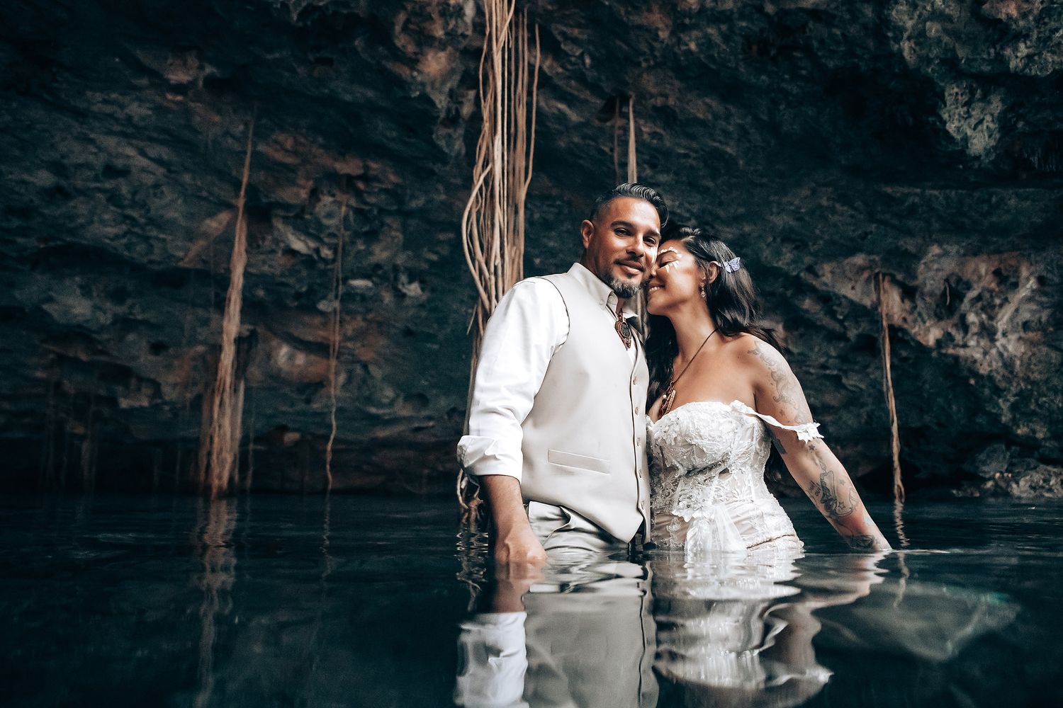A couple shares a tender moment in a cave cenote with natural vines and atmospheric lighting.