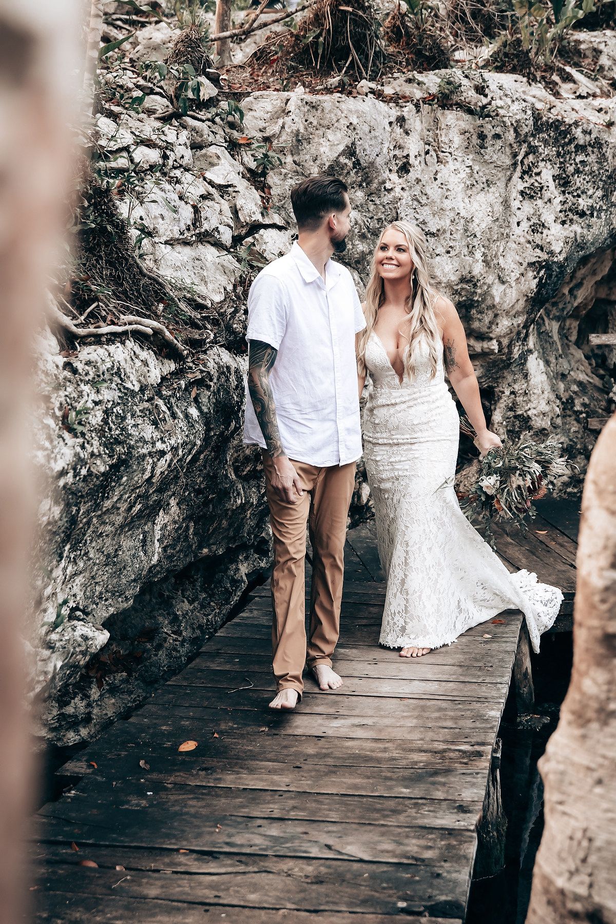 A couple walks together on a wooden boardwalk against a rocky cliff backdrop during a wedding photoshoot.