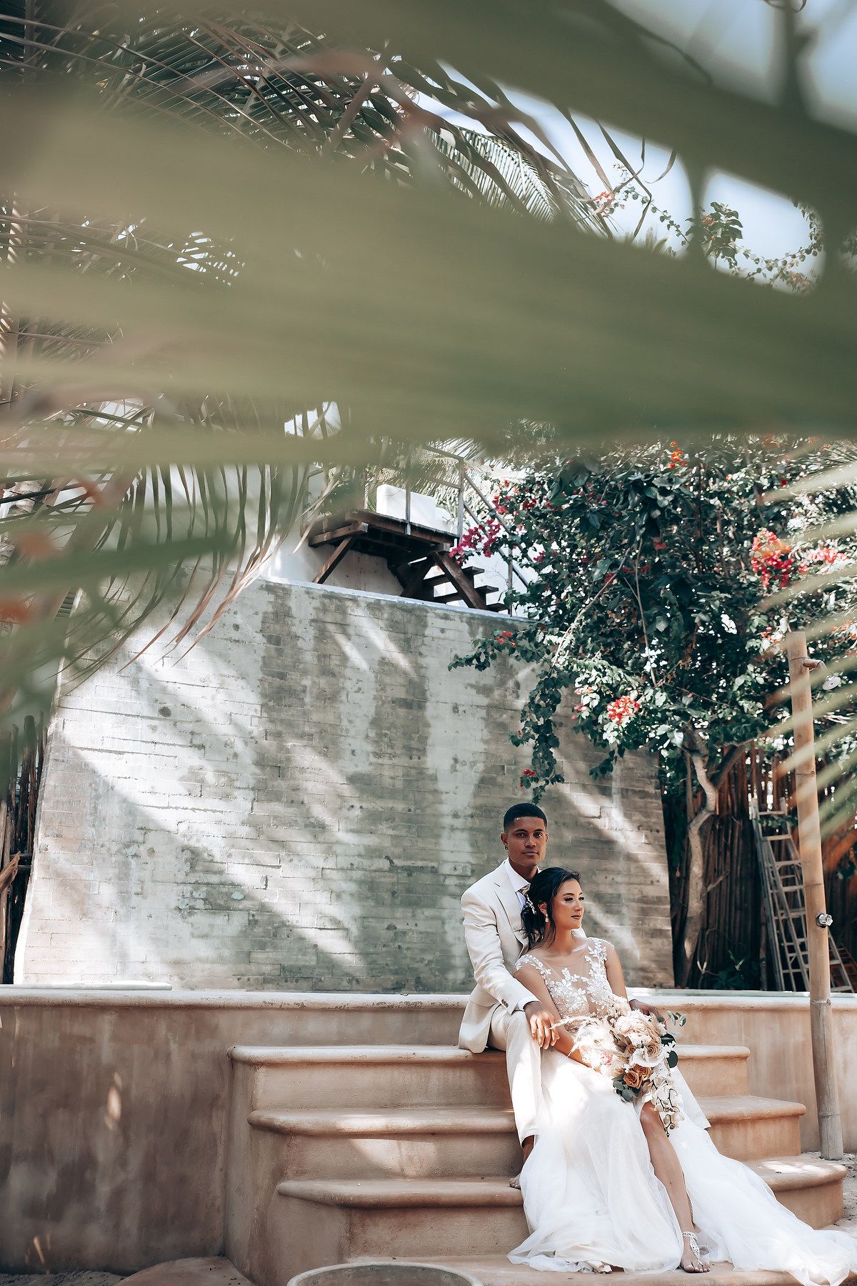 Newlyweds sitting together on garden steps framed by palm leaves and greenery.