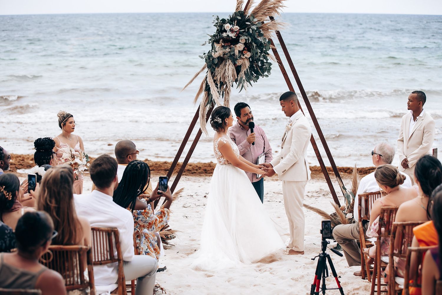 Beach wedding ceremony setup with wooden chairs and floral arch against ocean backdrop.
