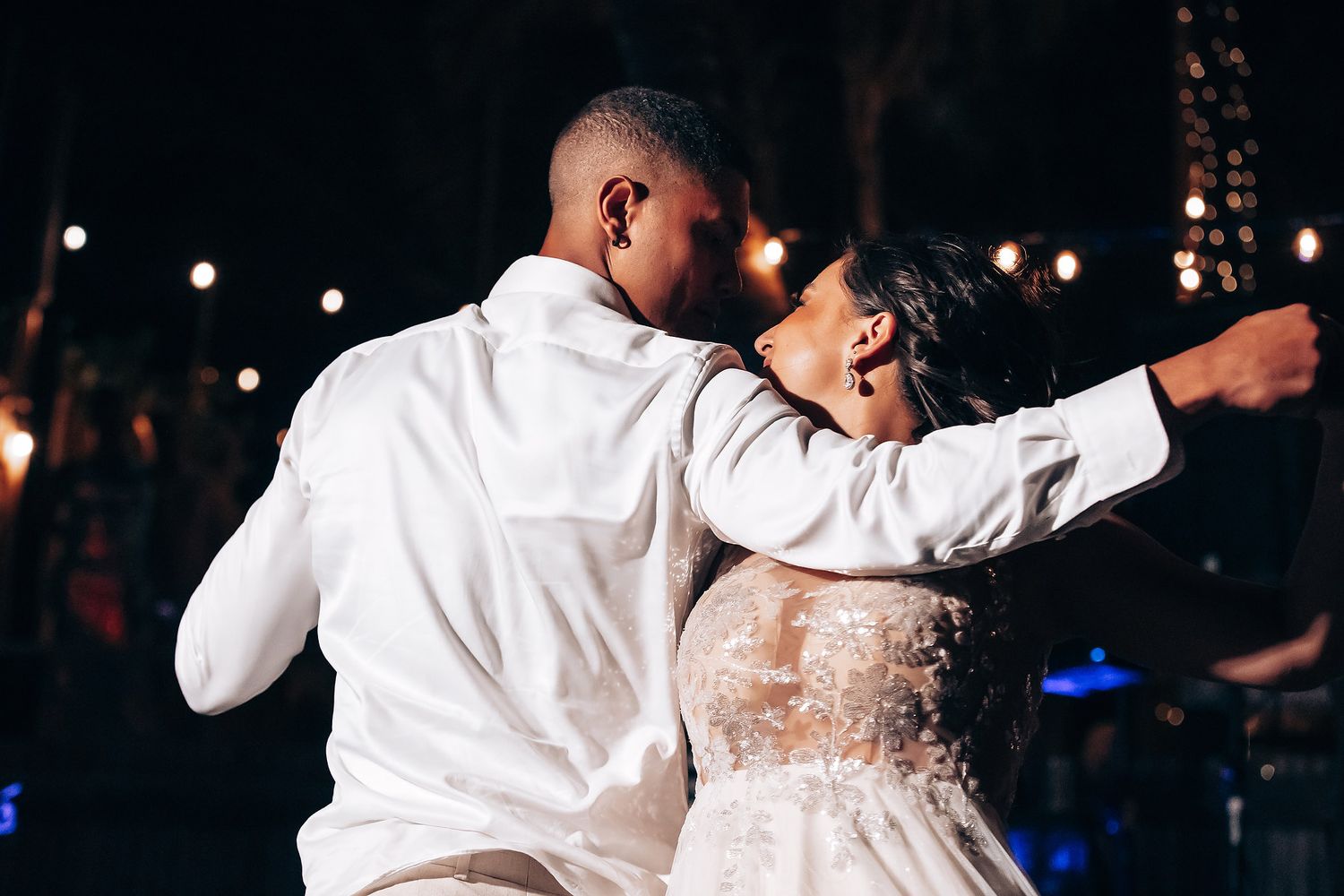 A couple shares an intimate dance moment at a wedding reception with romantic lighting and string lights overhead.
