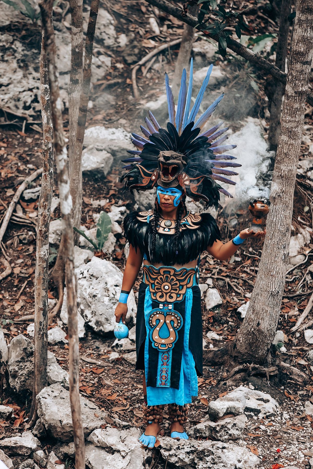 A person in traditional Aztec ceremonial dress with feathered headdress and turquoise clothing performs a ritual dance in nature.