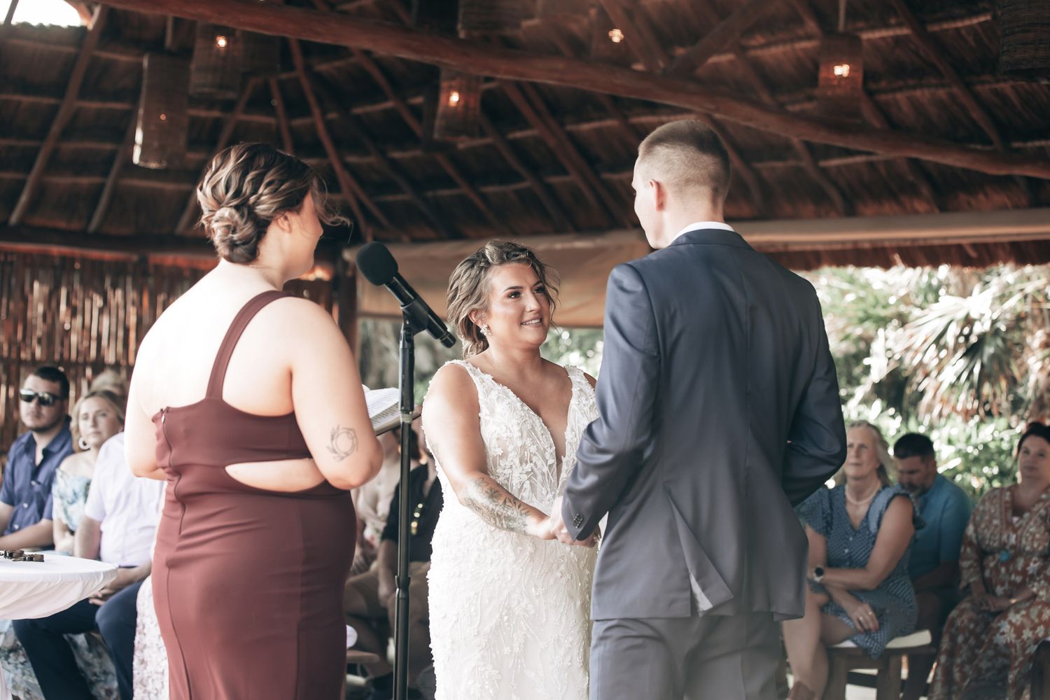 Bride and groom holding hands at rustic outdoor wedding ceremony while officiant reads vows at microphone.