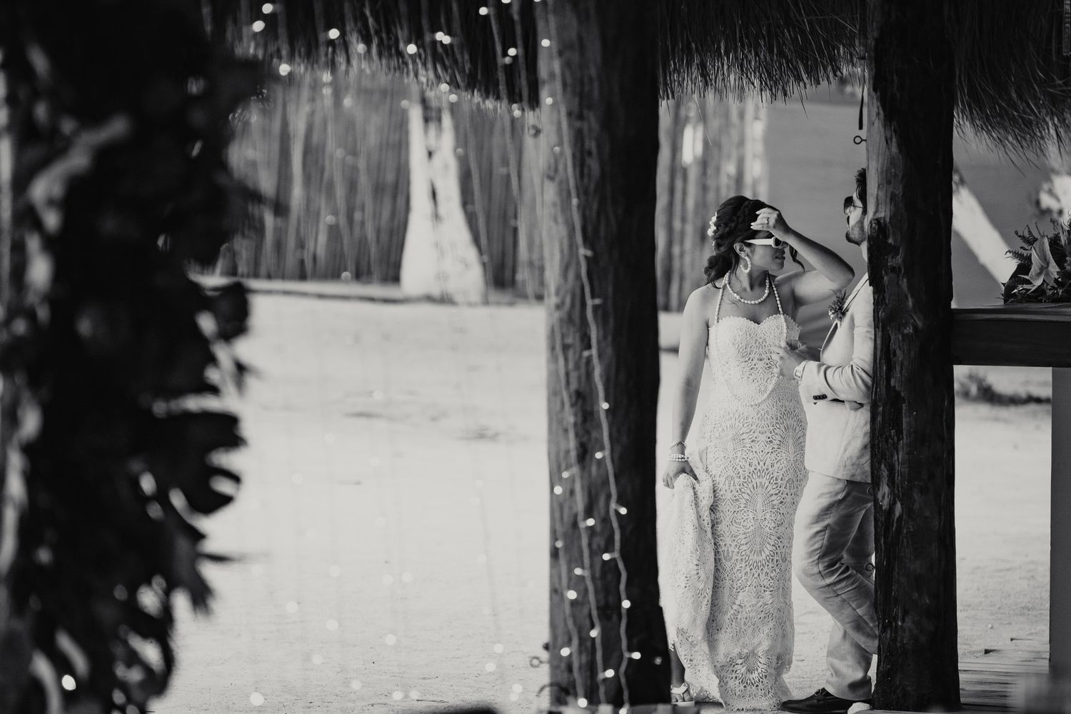 Bride in sparkly gown stands between palm trees with string lights at beach wedding.