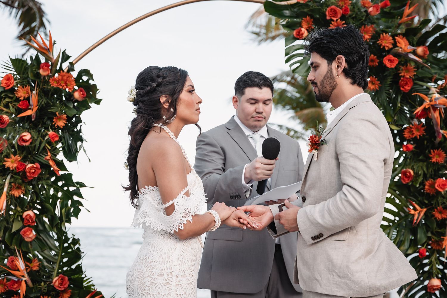 Couple exchanges rings at tropical beach wedding altar adorned with orange flowers and monstera leaves.